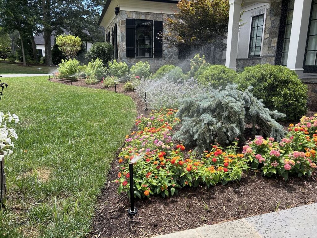 A lawn sprinkler system activated over a garden of flowers and shrubs.