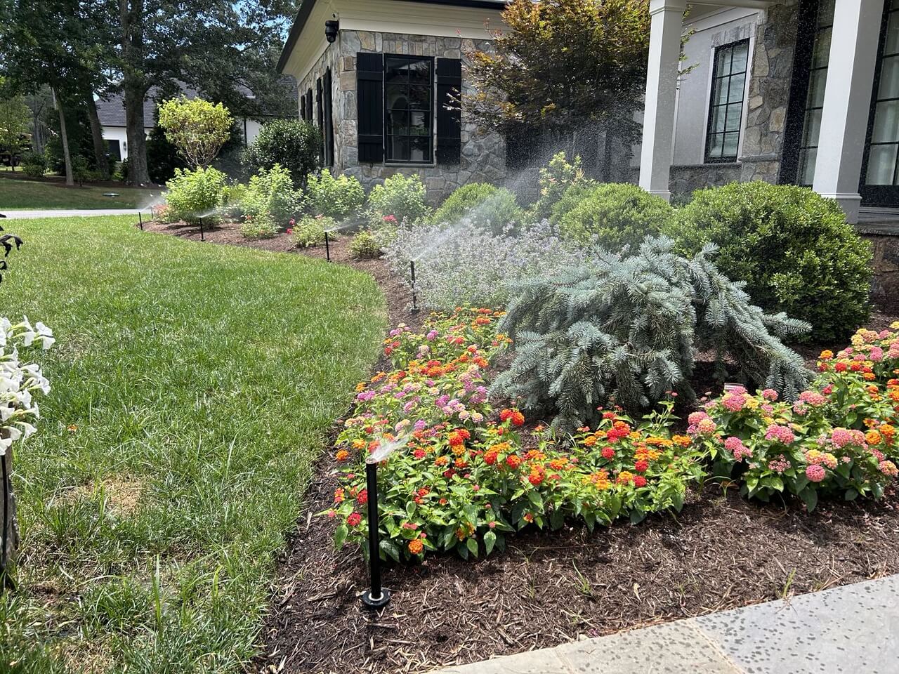 A lawn sprinkler system activated over a garden of flowers and shrubs.